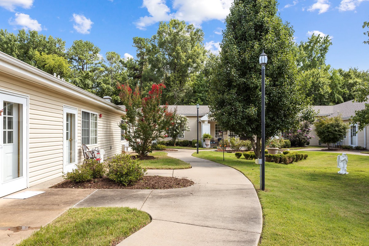 A scenic outdoor pathway bordered by grass and trees, leading past a building and a lamp post.