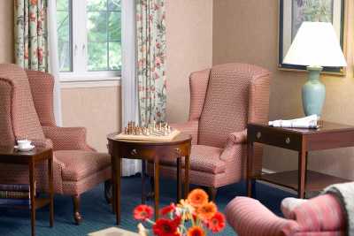 A cozy living room corner with armchairs, a chessboard on a side table, and a window with floral curtains.