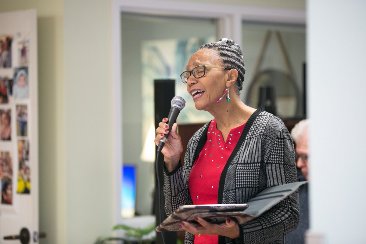 A woman wearing glasses, holding a microphone and a book, singing at an indoor event.