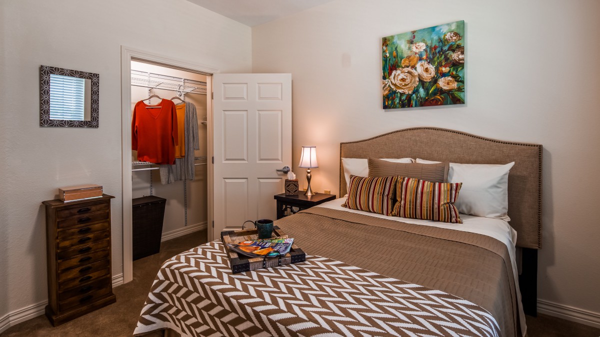 A cozy bedroom featuring a neatly made bed with striped pillows and patterned bedding, an open closet displaying clothes, and decorative elements like a painting and lamp.