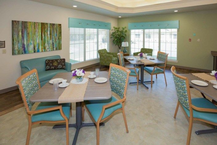 A cozy dining area with tables and chairs, featuring large windows and indoor plants.