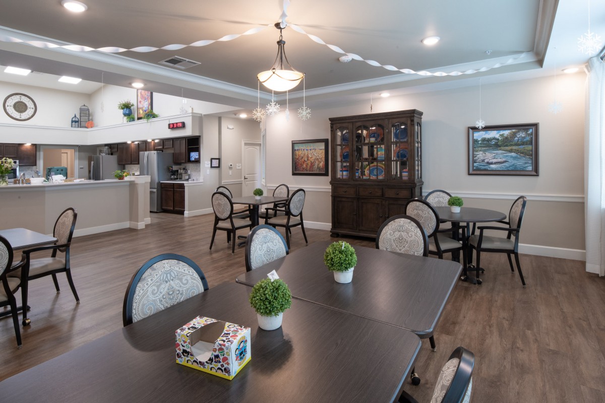 A cozy dining area with elegant decor, featuring multiple tables and chairs, a chandelier, and potted plants on wooden flooring.