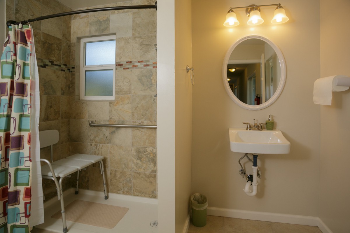 An accessible bathroom featuring a walk-in shower with a grab bar and a bench, alongside a wall-mounted sink and oval mirror under warm lighting.