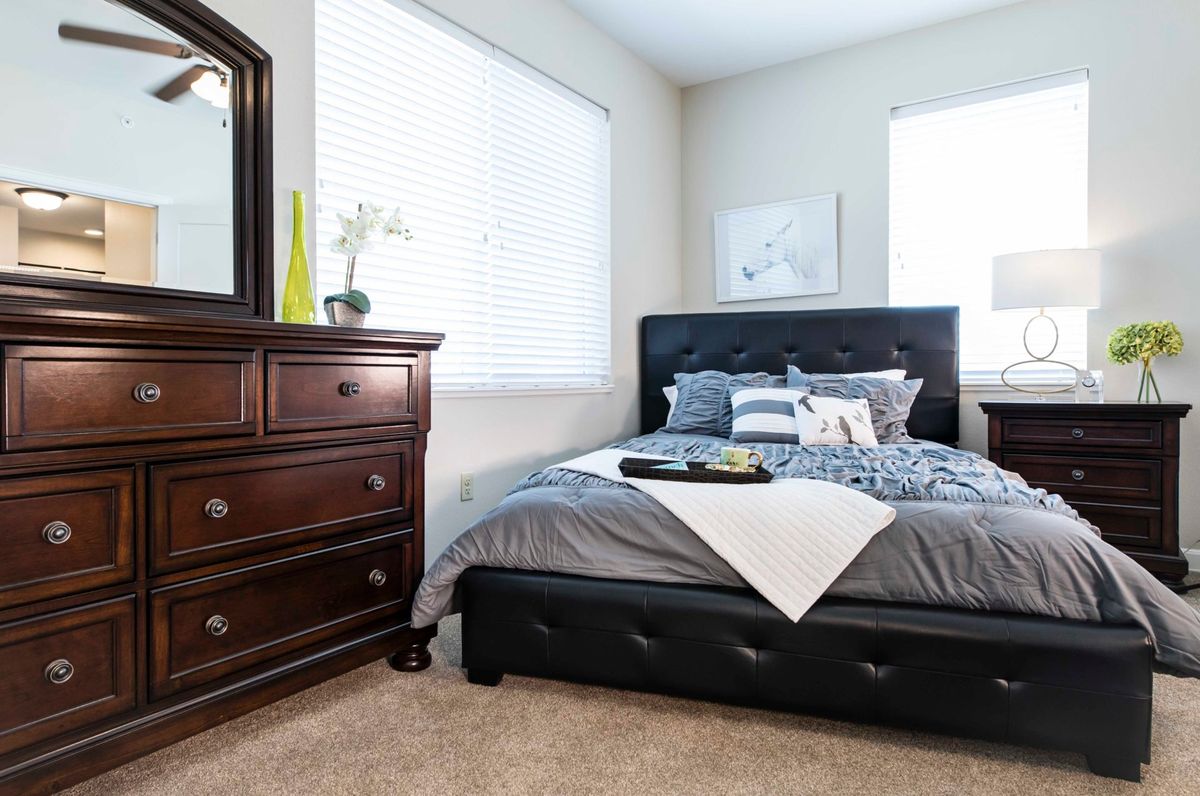 A modern and cozy bedroom featuring a dark leather bed frame, neatly arranged bed linens, a wooden dresser with a mirror, and minimalist decor.