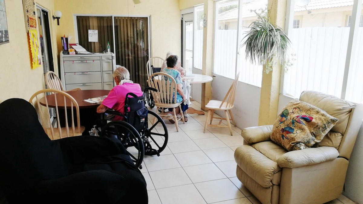 A cozy living area in a nursing home with elderly residents seated at tables and in wheelchairs.