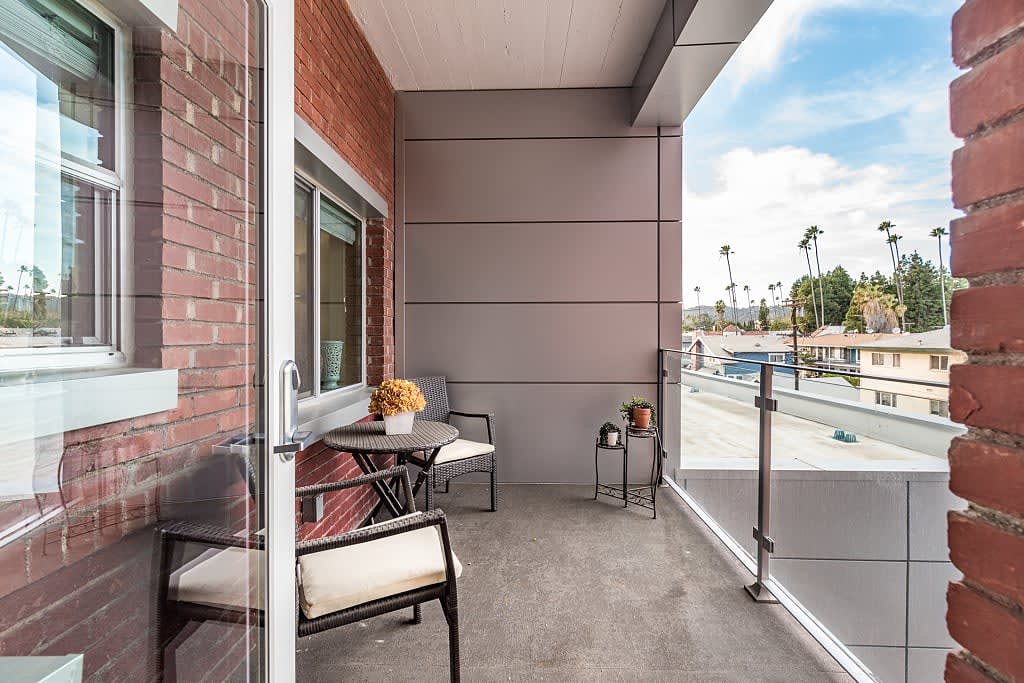 A modern balcony featuring comfortable seating and a glass railing, overlooking a street with palm trees.