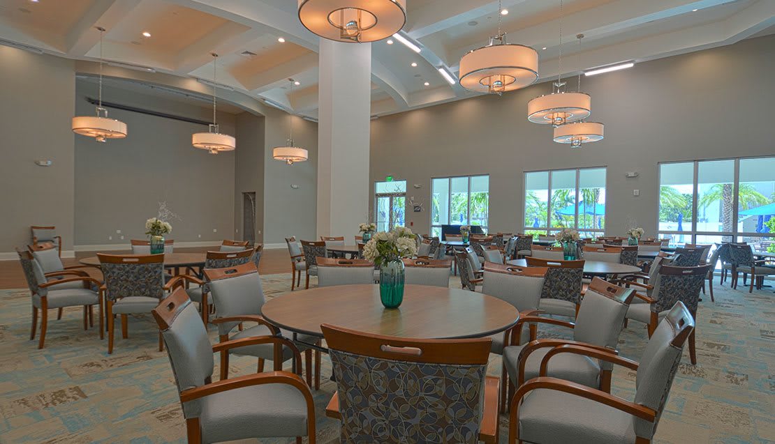 A spacious dining room with round tables and elegant chandeliers, decorated with flower arrangements.