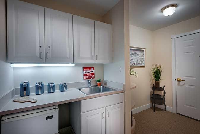 A compact kitchenette with white cabinets and a small sink, adjacent to a cozy hallway adorned with artwork and a potted plant.