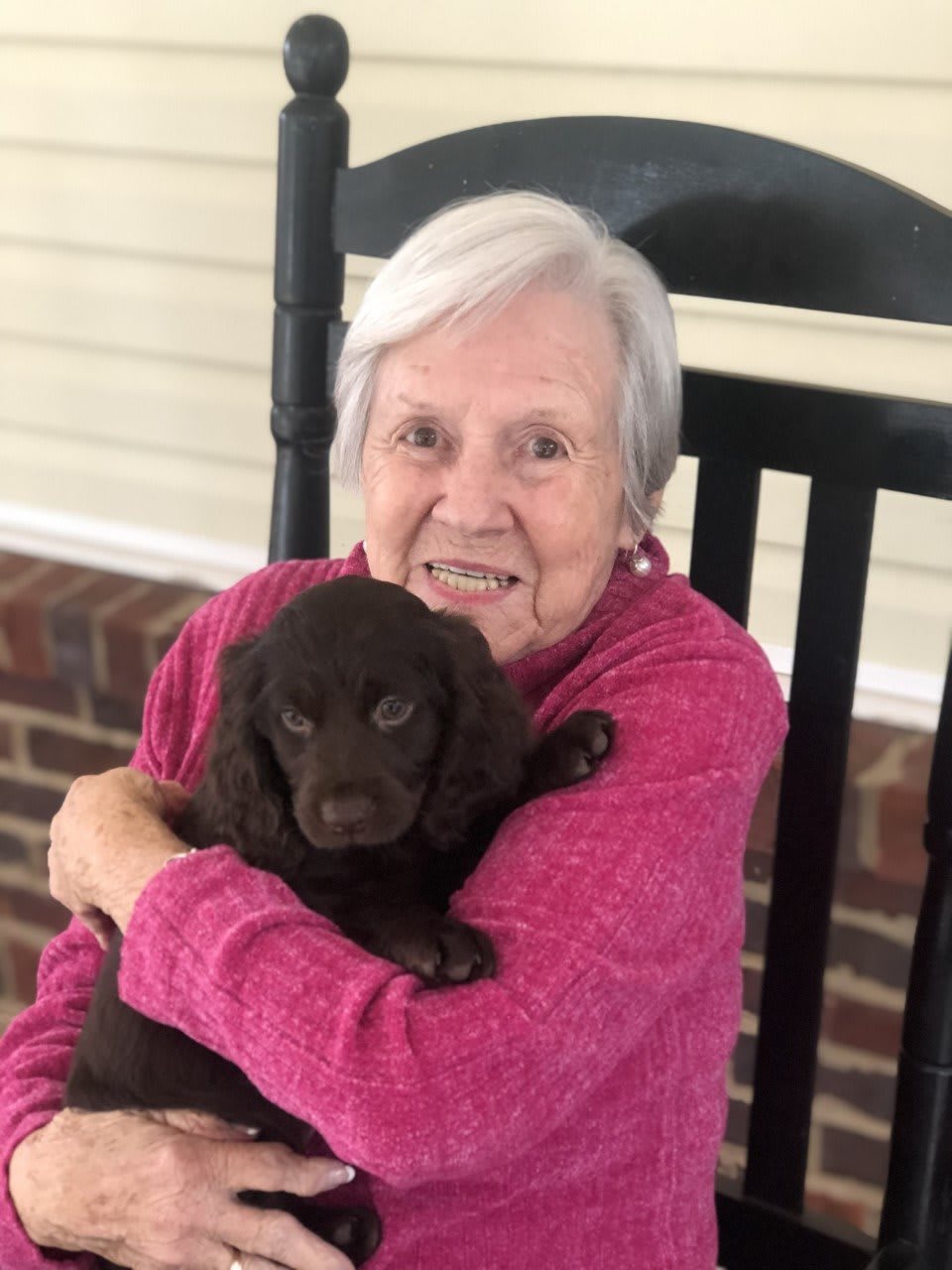 An elderly woman sitting on a porch in a rocking chair, holding and smiling at a puppy.