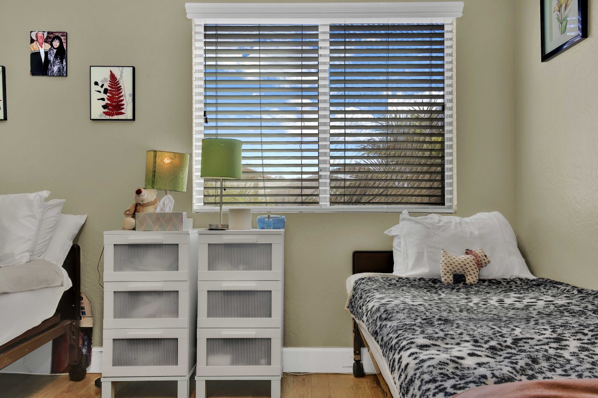 A cozy bedroom featuring a bed with patterned bedding, a nightstand with lamps, and a large window with blinds.