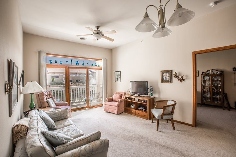 A cozy living room with a sofa, armchair, television, and ceiling fan, featuring large windows that let in natural light.