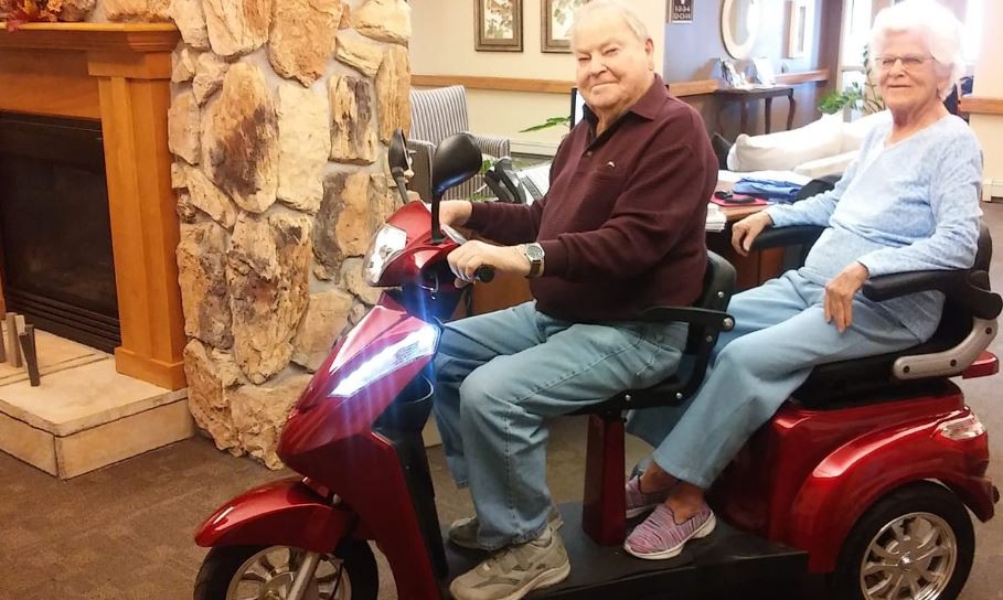 An elderly person smiling while sitting on a scooter indoors.