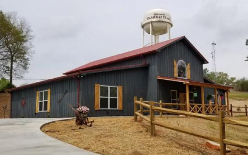 A rural barn with a red roof and adjacent water tower, surrounded by farm equipment and a wooden fence.