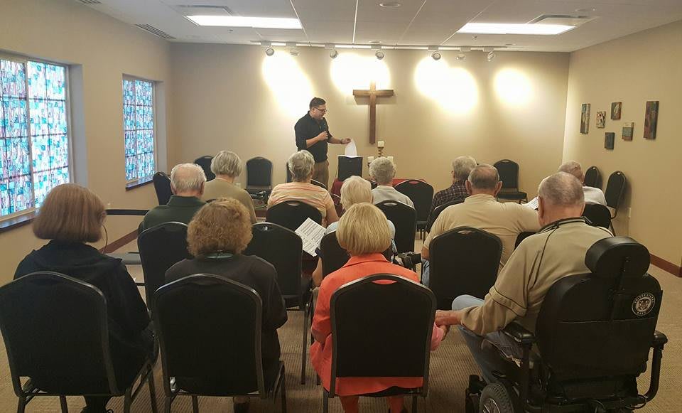 A speaker presents to an audience in a room with a cross on the wall.