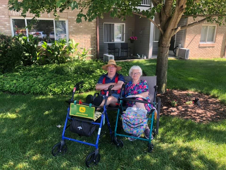 Two elderly individuals sitting outside in wheelchairs, enjoying a sunny day in the garden.