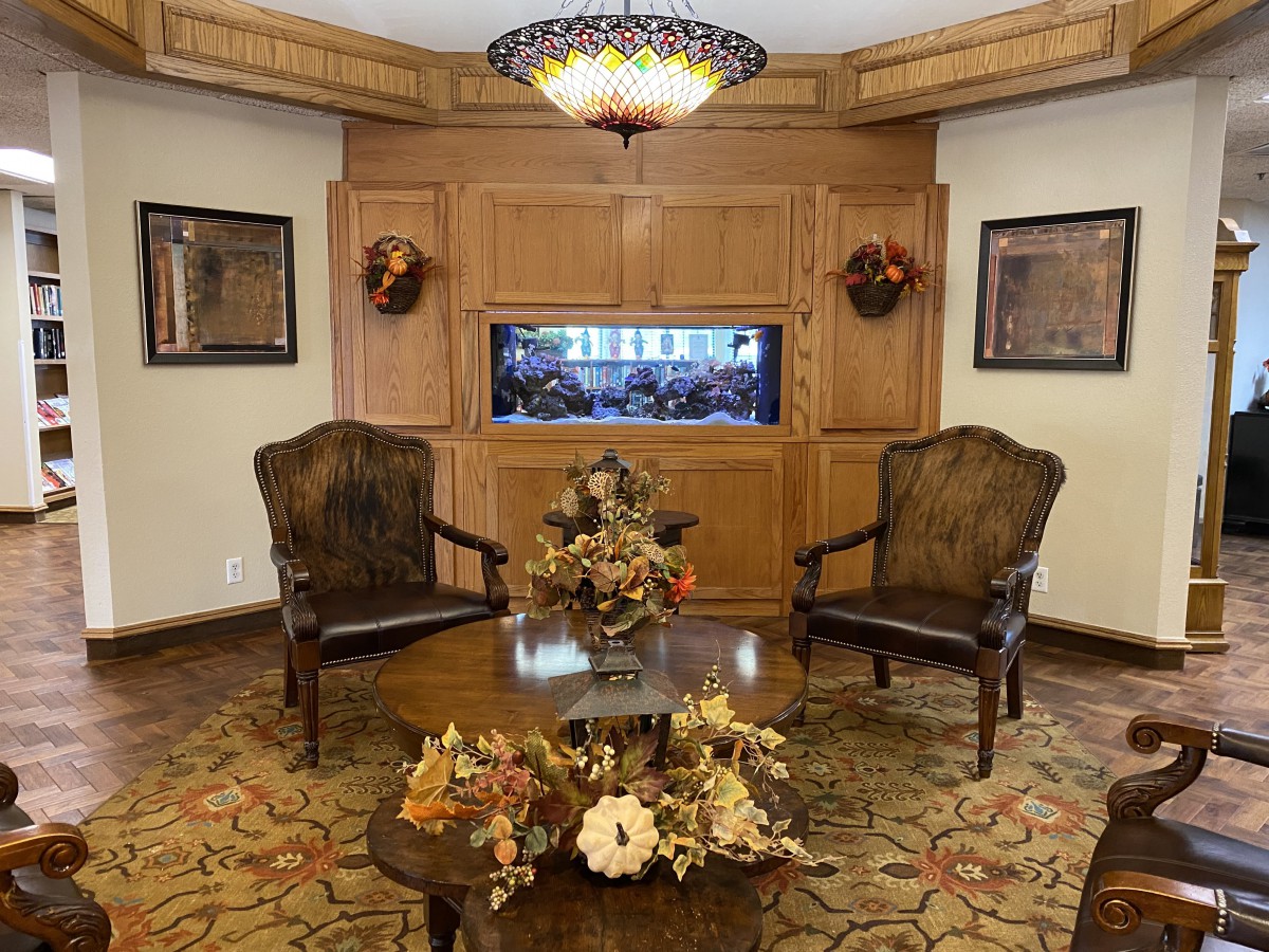 A cozy interior setting featuring two armchairs, a wooden table with floral arrangements, and an aquarium set in wood-paneled walls beneath a decorative chandelier.