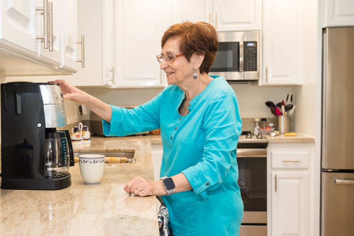 A woman in a blue shirt making coffee in a bright kitchen.