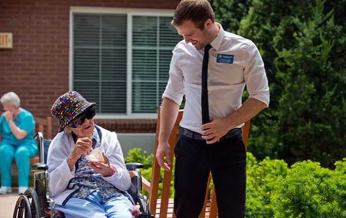 A caregiver interacting with an elderly person seated in a wheelchair outdoors.
