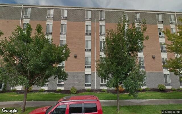 A multi-story apartment building with a brick facade, surrounded by trees and a red car parked in front.