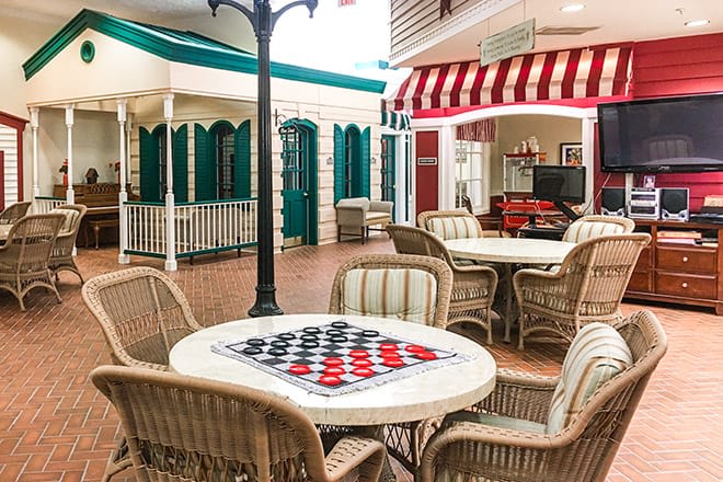 An inviting indoor space with tables and chairs, featuring a checkerboard for games, surrounded by decorative storefronts.