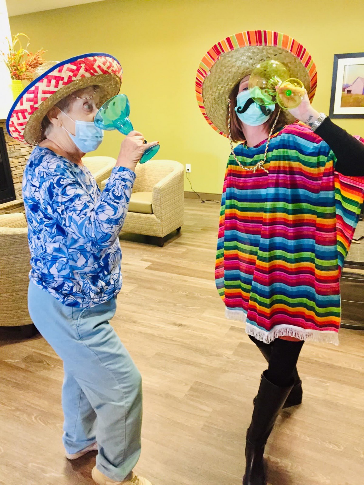 Two people dressed in festive attire, wearing sombreros and masks, pose playfully indoors.
