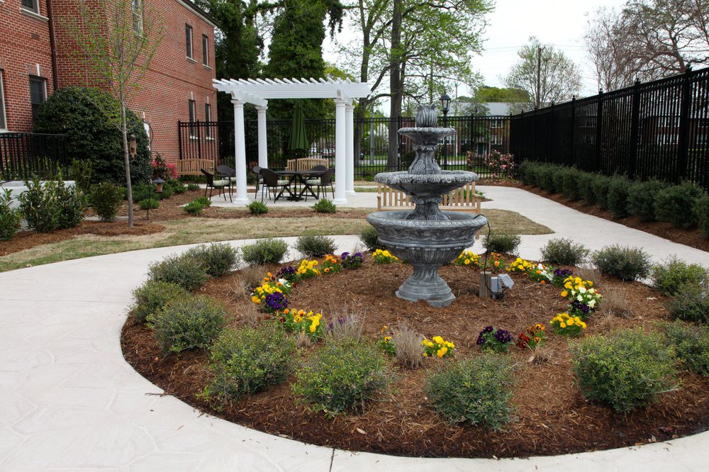 A neatly landscaped garden featuring a central fountain and a pergola in the background, surrounded by vibrant flowers and pathways.