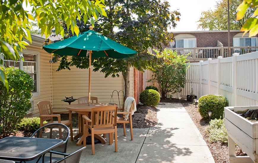 A sunny backyard with outdoor seating and a green umbrella, surrounded by plants.