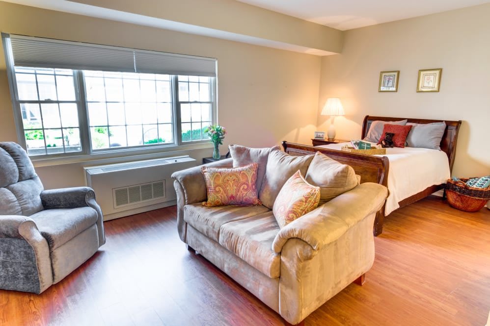 A cozy living room with a sofa and armchair in front of a large window, featuring warm lighting and wooden flooring.