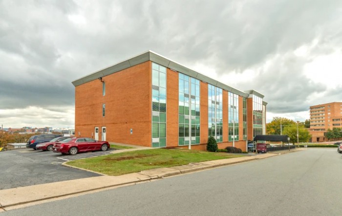 A modern brick office building with large glass windows and a parking lot under an overcast sky.