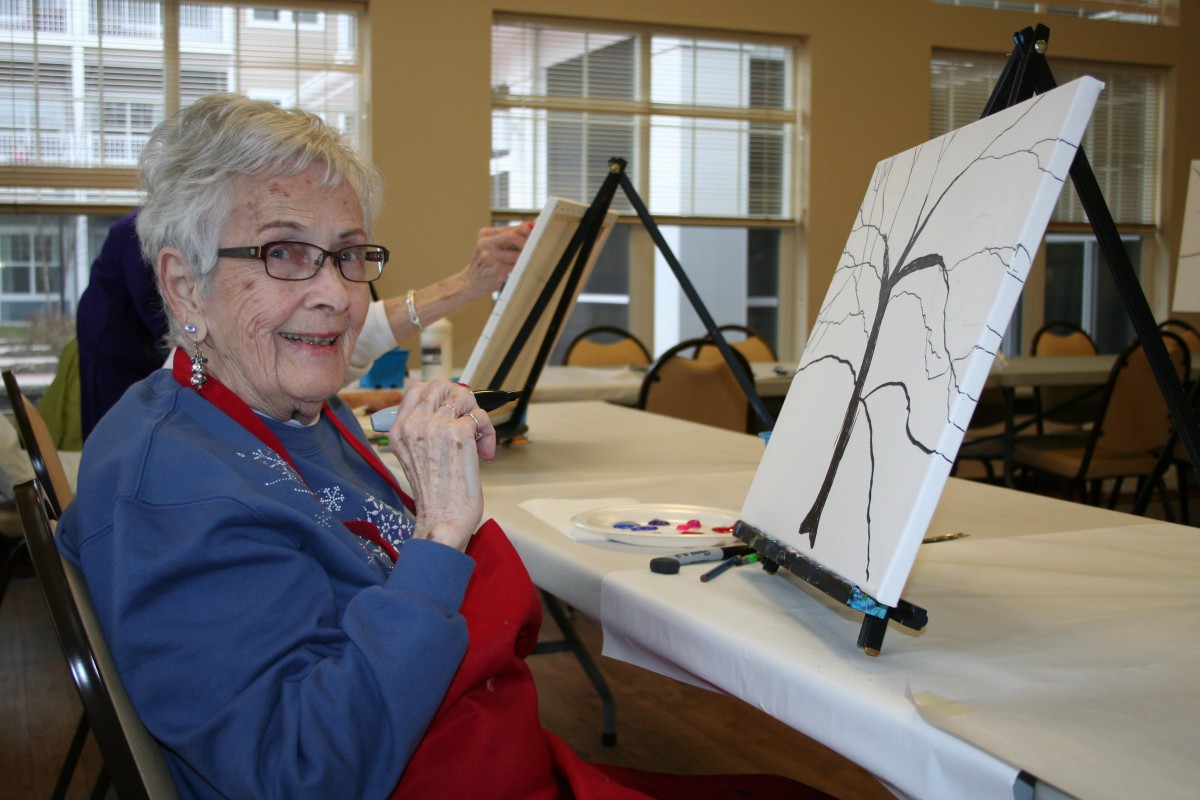 An elderly woman smiling while painting on a canvas during an art class.