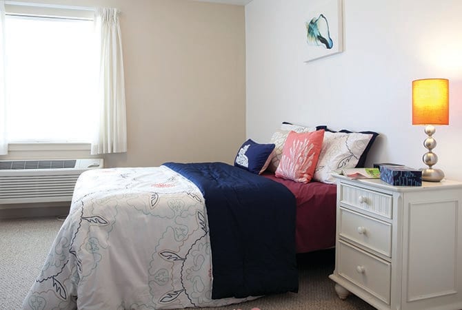 A cozy bedroom with a neatly arranged bed, featuring colorful pillows and a white dresser.