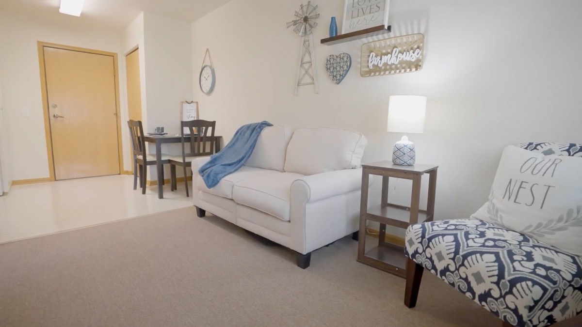 A cozy living room with a white sofa, wall decorations, and a small dining area in the background.