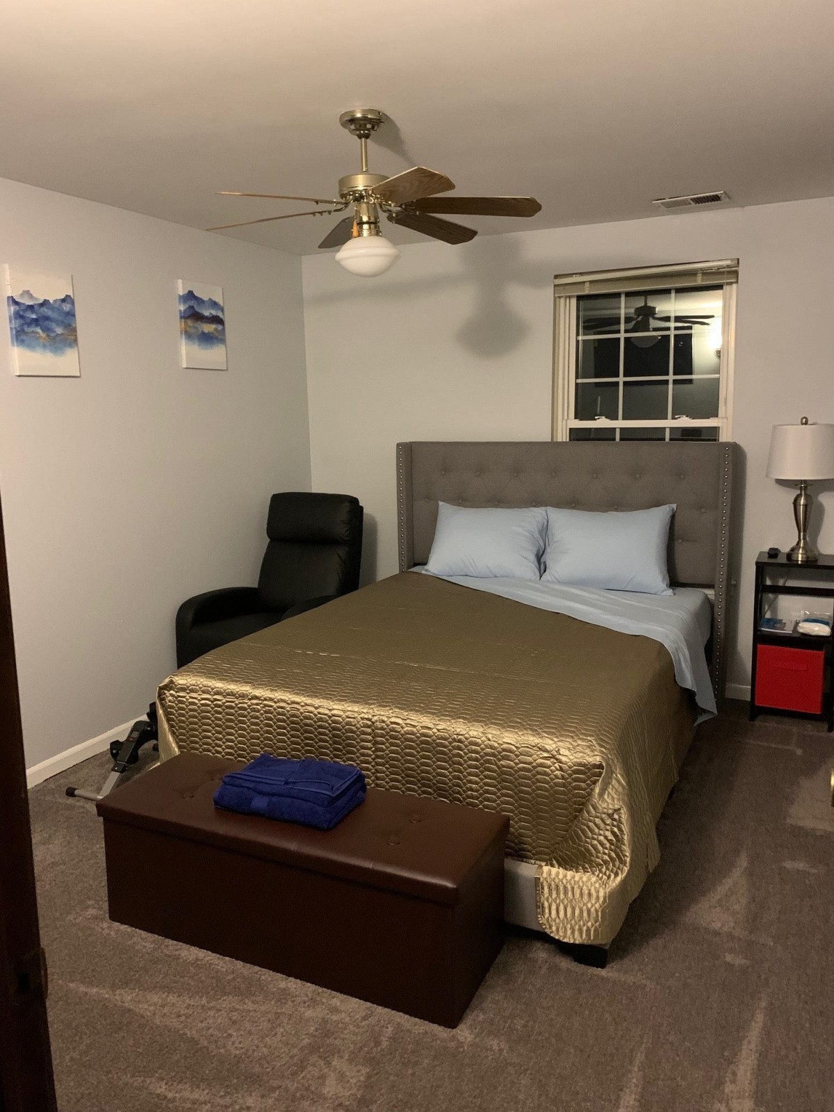 A neatly arranged bedroom featuring a bed with blue pillows and gold bedding, a black chair, nightstand with lamp, and ceiling fan.