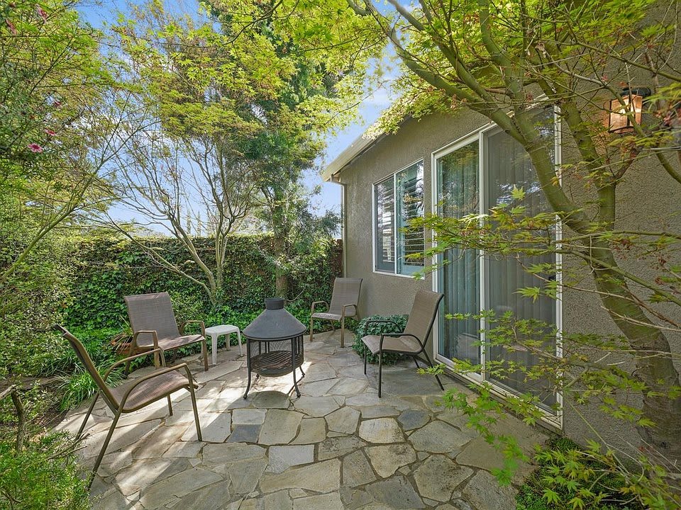 A cozy patio area with seating around a fire pit, surrounded by lush greenery and part of a house exterior.