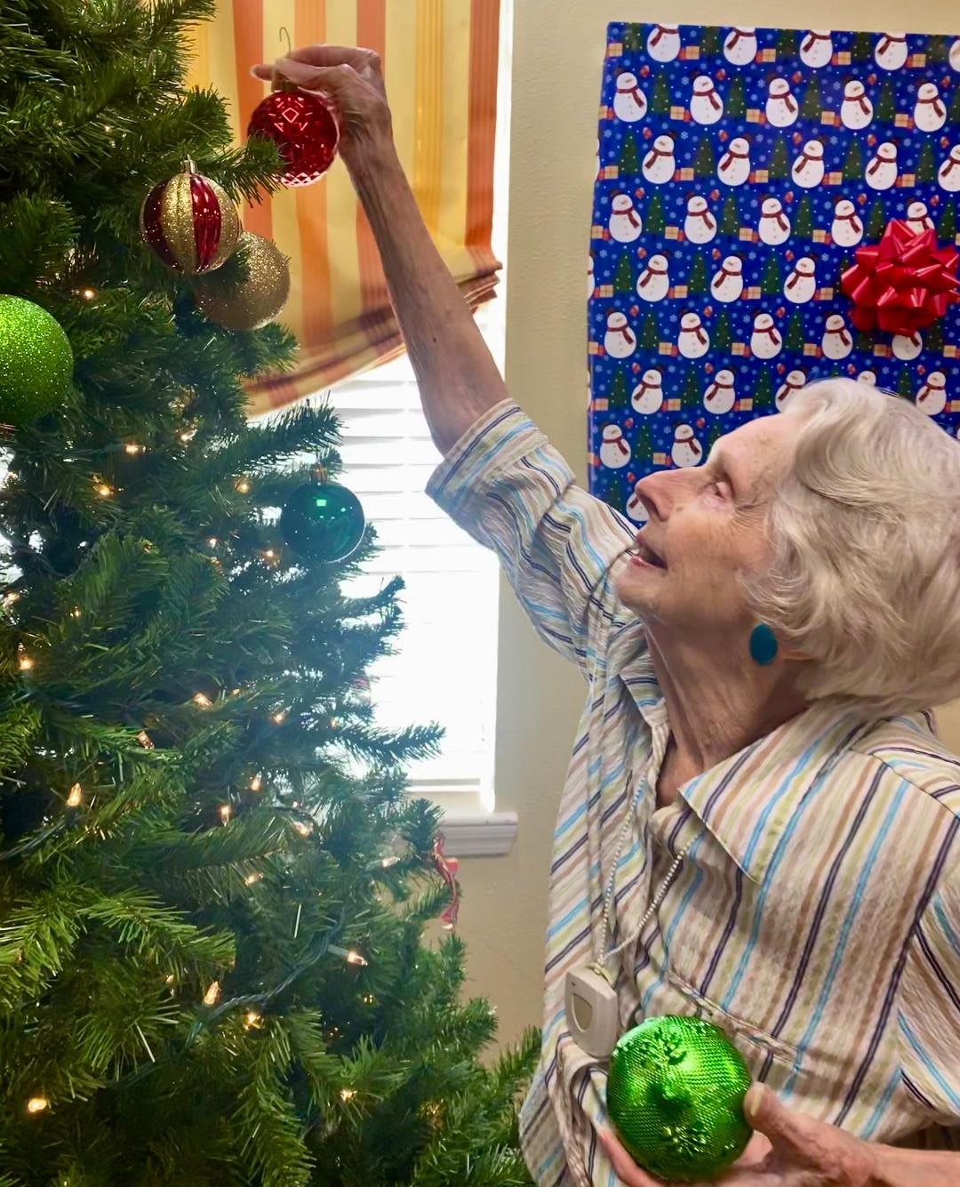 An elderly woman joyfully decorating a Christmas tree with ornaments.