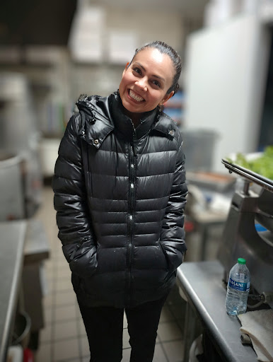 A cheerful woman wearing a black puffer jacket stands in a kitchen, radiating positivity.