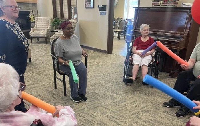 A group of senior citizens participating in a seated exercise session indoors, using pool noodles.