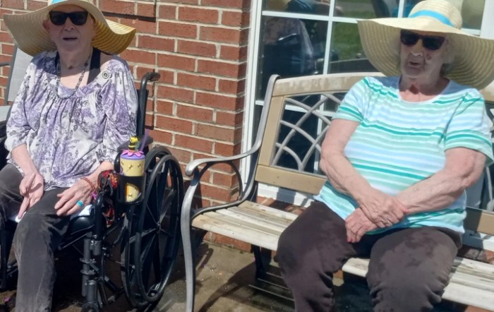 Two elderly individuals sitting outdoors, one in a wheelchair and the other on a bench, both wearing hats.