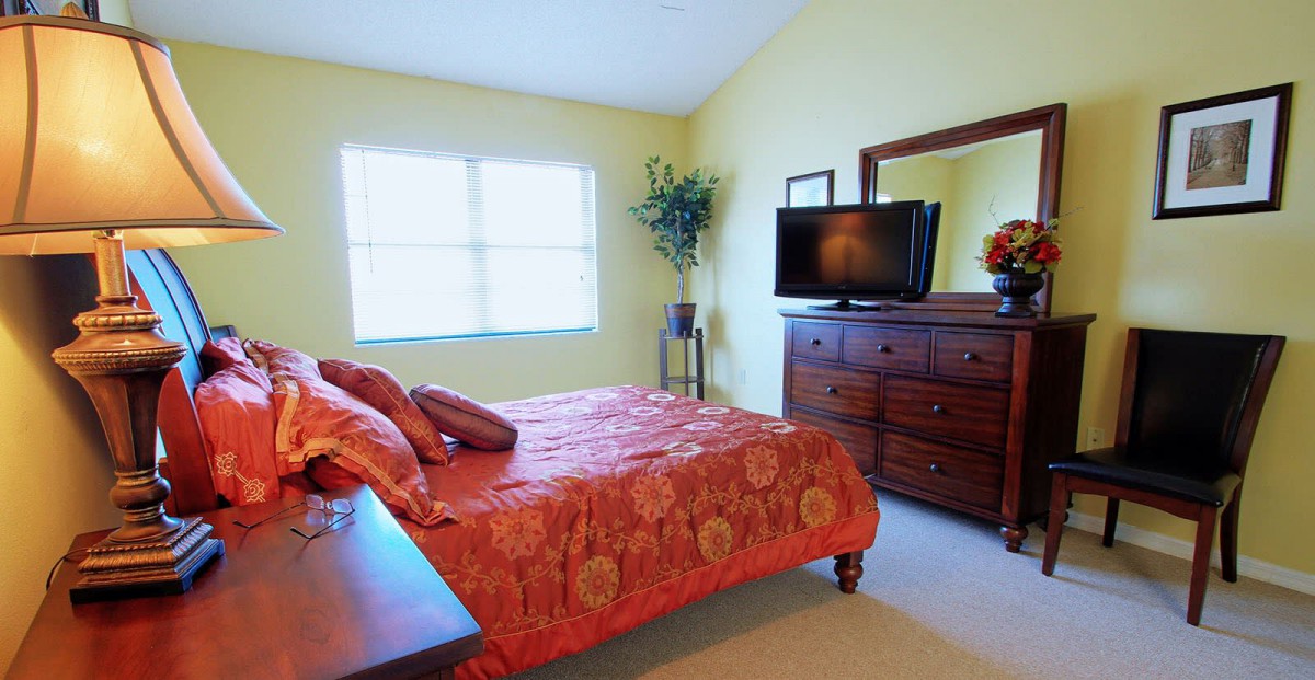 A cozy bedroom with orange bedding and a wooden dresser, featuring a television and window.