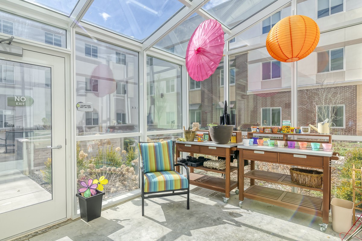 A bright sunroom with a glass ceiling, featuring colorful hanging decorations, a striped chair, and a variety of potted plants.