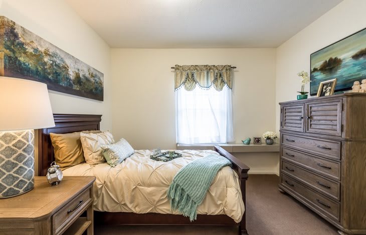 A cozy bedroom with a wooden bed, bedside table, and a tall dresser next to a window.
