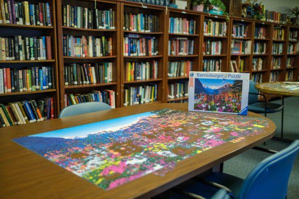 A completed jigsaw puzzle depicting colorful scenery is displayed on a table in a library, surrounded by shelves full of books.
