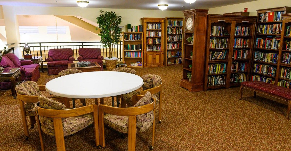 A cozy library reading area with bookshelves, a round table, and comfortable chairs.
