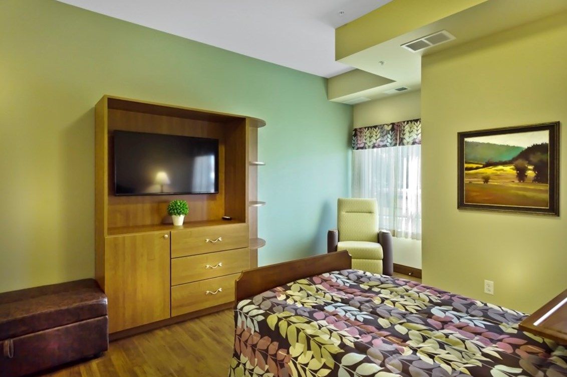 A well-furnished hospital room featuring a bed with floral bedding, a wooden cabinet with a television, and an armchair by the window.