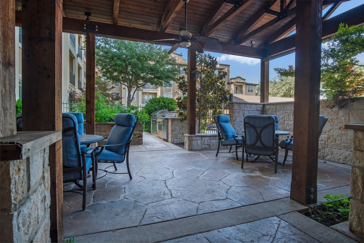A cozy outdoor patio with seating under a covered pavilion, surrounded by greenery and buildings.