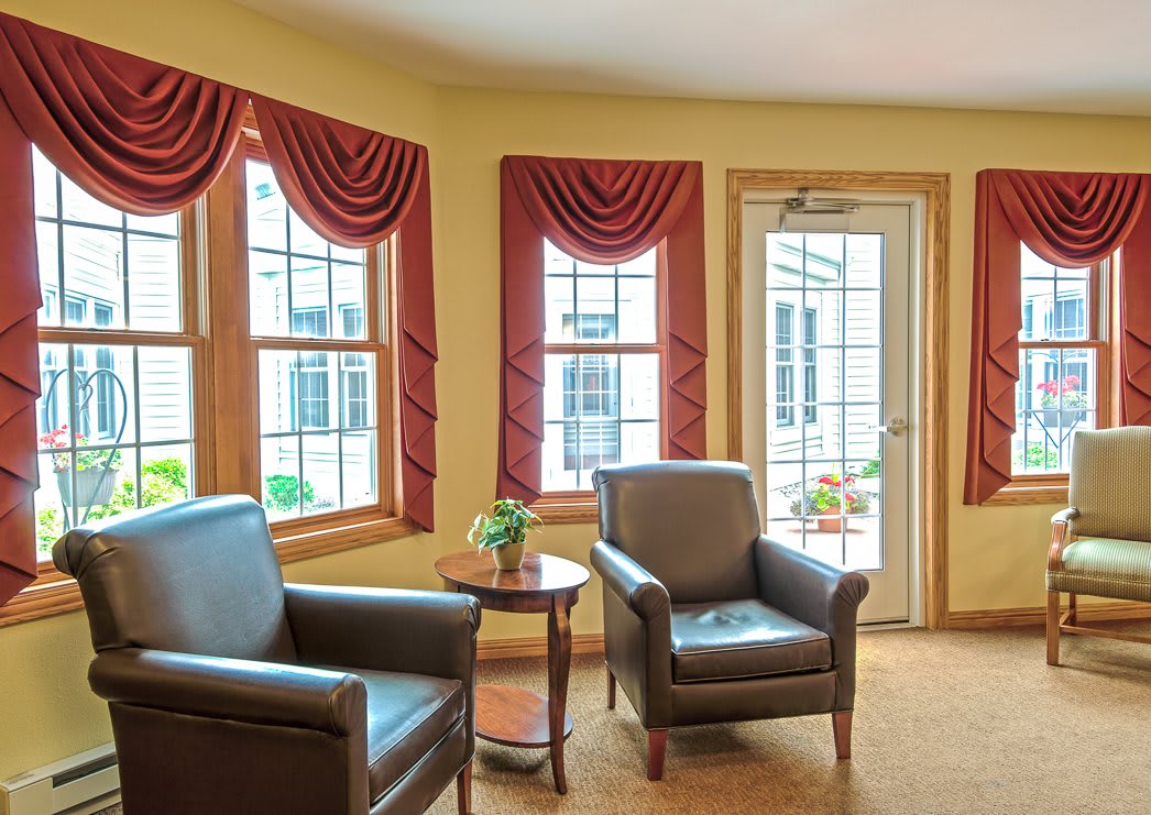 A cozy living room with two leather chairs, a small table, and large windows adorned with red curtains.
