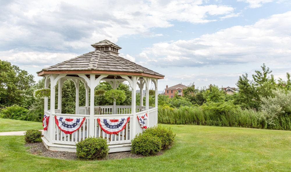 A white gazebo decorated with red, white, and blue bunting sits on a lush green lawn under a partly cloudy sky.