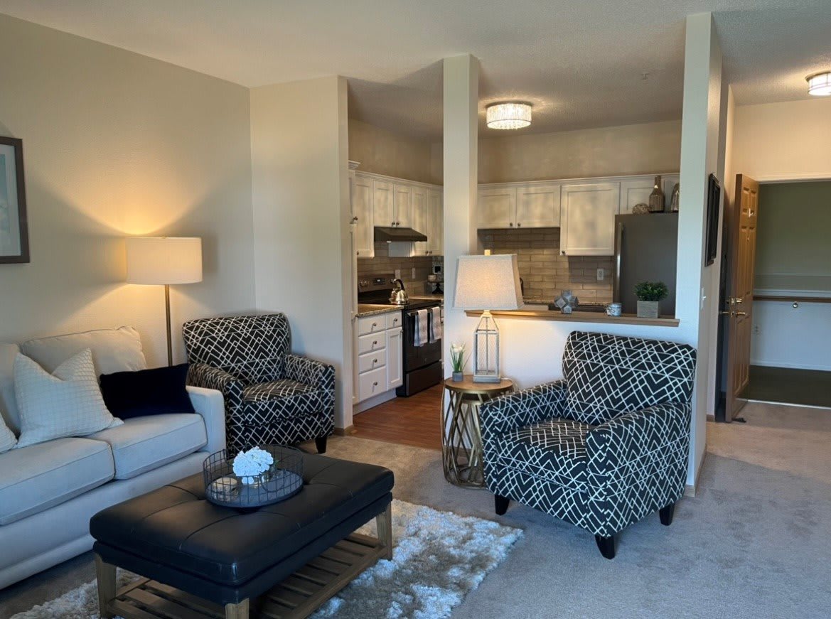 A modern apartment living room with an open kitchen layout, featuring patterned armchairs and ambient lighting.