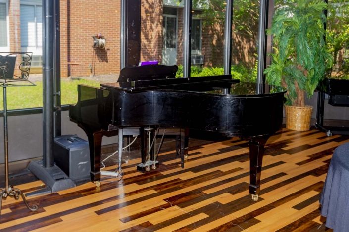 A grand piano positioned in a sunlit room with large windows and a polished hardwood floor.