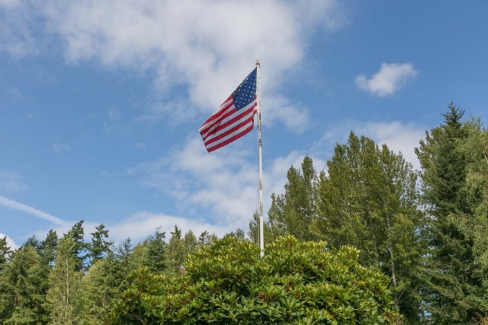 An American flag waving atop a flagpole surrounded by lush trees under a partly cloudy sky.